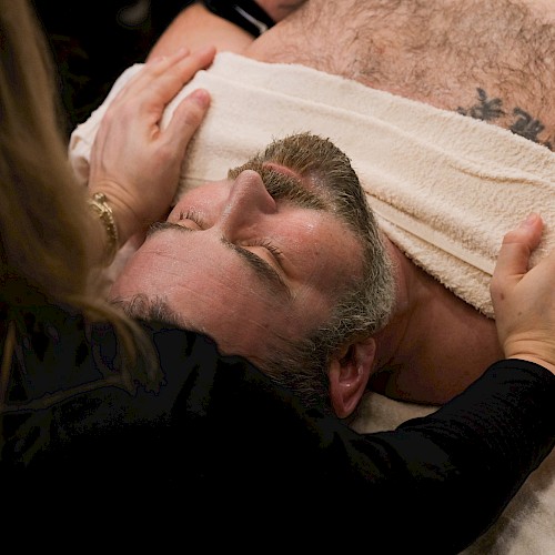 A woman comforting a man lying on a massage table, gently holding his head and shoulders.