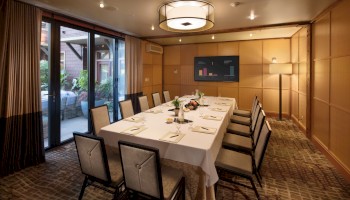 A formal dining room with a long, L-shaped table set for twelve, white tablecloths, elegant place settings, a wall TV, warm lighting, and glass doors opening to an outdoor patio.