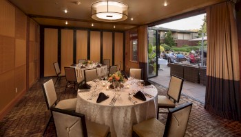 A fine dining room set for a formal event with round tables, white tablecloths, folded napkins, candles, and chairs; open doors reveal an outdoor patio.