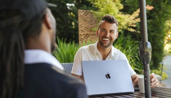 Two people at an outdoor table; a man with a laptop (Apple logo) smiling at a person wearing a cap, in a sunny garden setting.