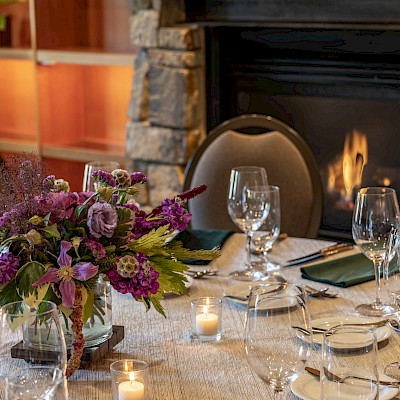A beautifully set dining table with a floral centerpiece, candles, wine glasses, and silverware in front of a fireplace.