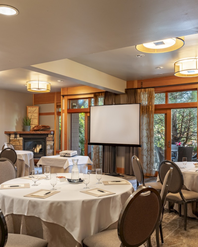 A cozy, elegantly set meeting or conference room with round tables, a projector, a screen, and a scenic outdoor view through large windows.