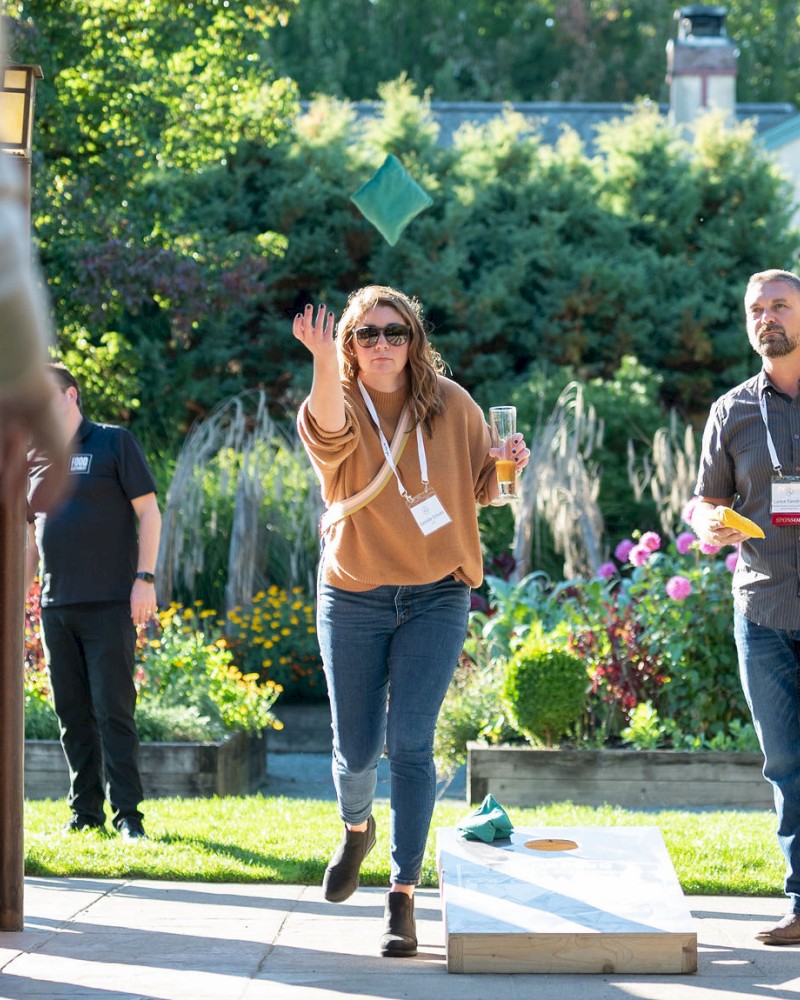 People outdoors playing cornhole—one woman throws a bag toward the board while others watch and chat in a sunny garden area.
