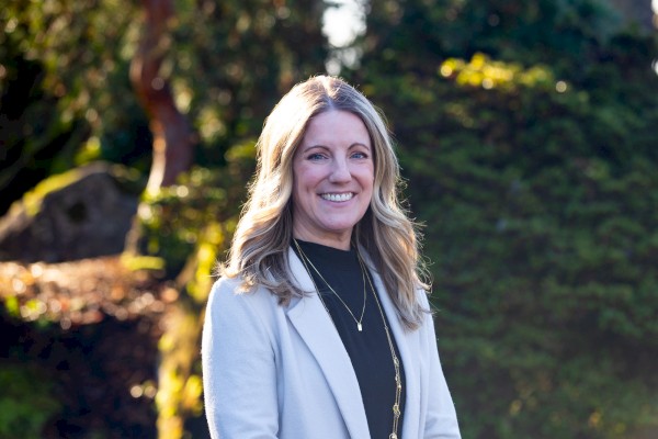 A smiling woman with blonde hair wearing a light blazer and black top stands outdoors, greenery and sunlight in the background.