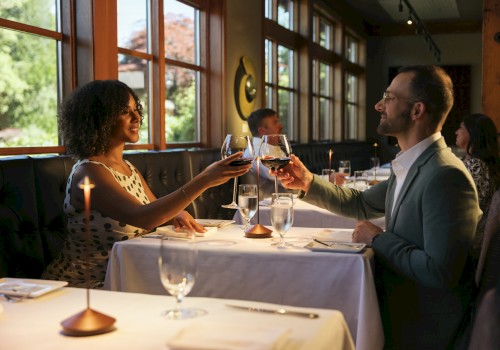 Two people toast at a candlelit restaurant table, smiling and clinking glasses, while other diners sit in a cozy, sunlit dining room.