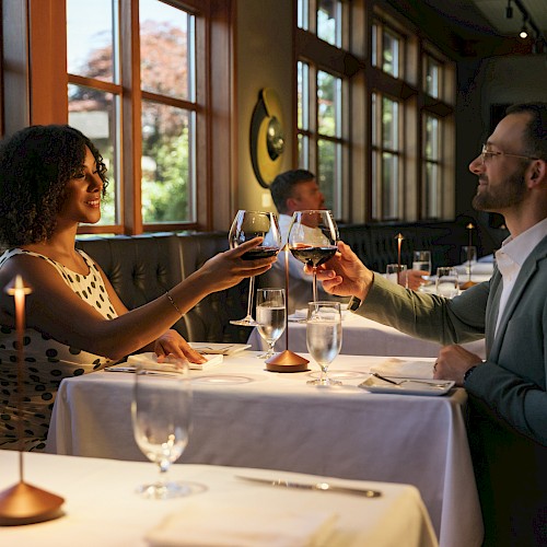 Two people toast at a candlelit restaurant table, smiling and clinking glasses, while other diners sit in a cozy, sunlit dining room.