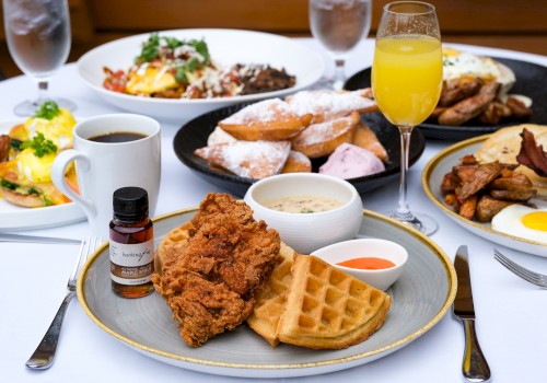 A table full of breakfast dishes: fried chicken with waffles, syrup, dipping sauces, coffee, orange juice, eggs, toast, pastries, and various plates in a bright restaurant setting.