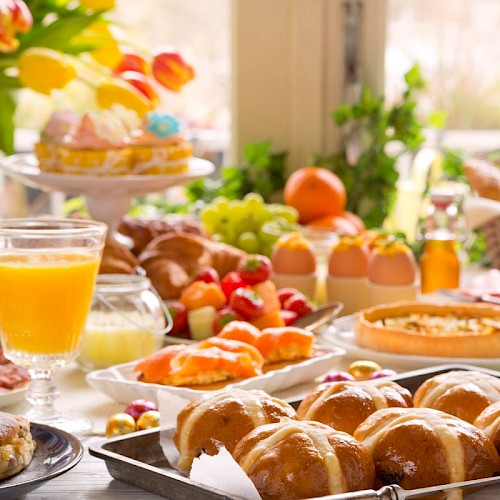 A colorful breakfast spread features pastries, fruits, eggs, juice, and baked goods on a sunny table.