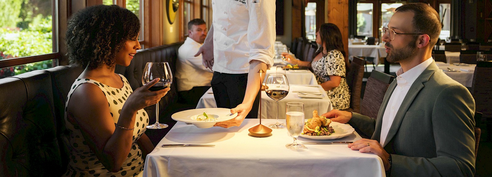 A woman in a polka dot dress enjoys wine with a man in a tuxedo at a restaurant, while a waiter serves them, surrounded by other diners.