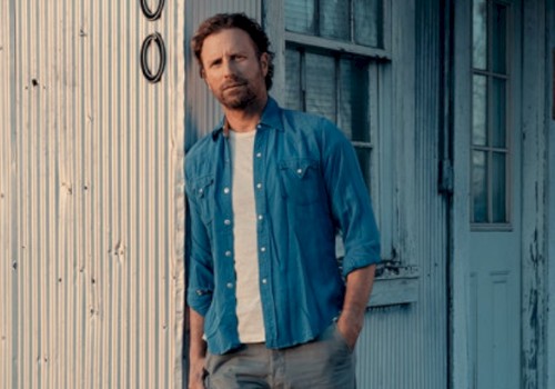 A man in a blue shirt stands against a rustic building with a corrugated metal wall and a window in the background.