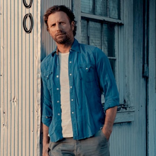A man in a blue shirt stands against a rustic building with a corrugated metal wall and a window in the background.
