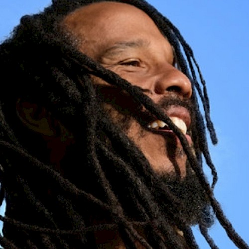 A person with long dreadlocks smiling against a blue sky background.
