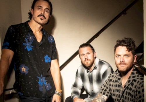 Three men are posing on and near a staircase in a casual setting, with a mix of floral and checkered shirts.