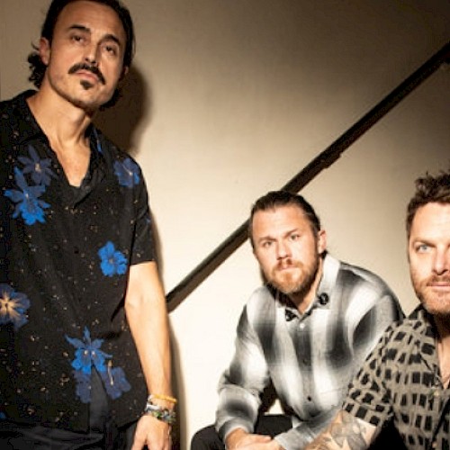 Three men are posing on and near a staircase in a casual setting, with a mix of floral and checkered shirts.