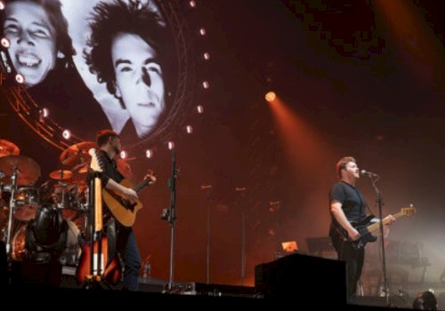 A band performs on stage with large portraits of two men in the background, illuminated by orange and white stage lights.