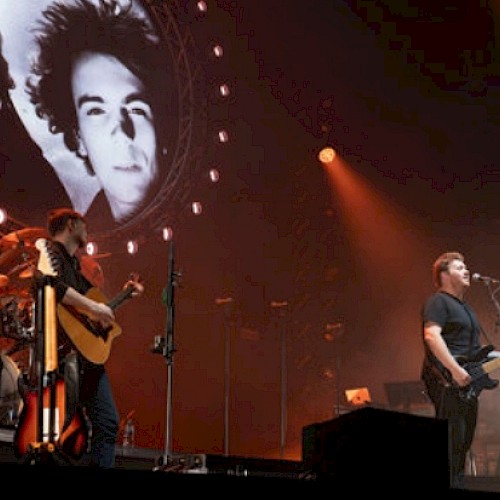 A band performs on stage with large portraits of two men in the background, illuminated by orange and white stage lights.