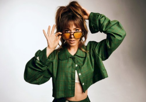A woman with brown hair in a green checked jacket, slightly lifting her glasses and gazing at the camera.