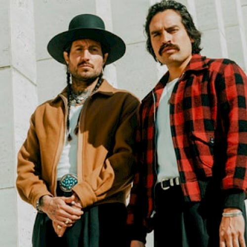 Two men with vintage style outfits pose outdoors, one with a wide-brimmed hat and the other with a mustache and checkered shirt.