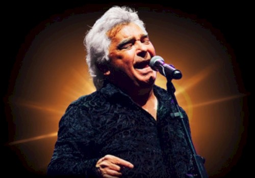 A man with white hair passionately sings or speaks into a microphone against a glowing background.