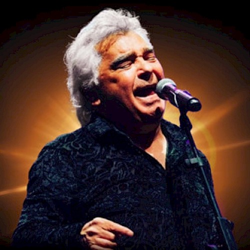 A man with white hair passionately sings or speaks into a microphone against a glowing background.