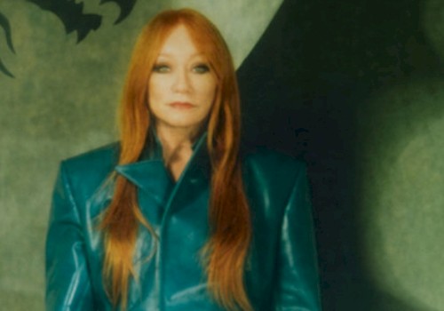 A woman with long red hair in a shiny teal jacket stands against a dark, artistic background.