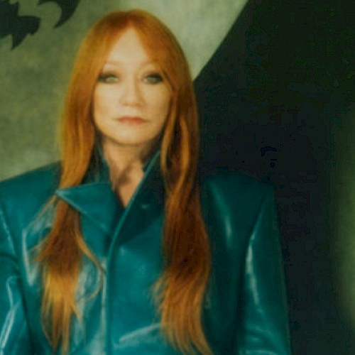 A woman with long red hair in a shiny teal jacket stands against a dark, artistic background.