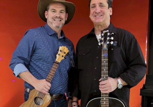 Two men with guitars, smiling on a stage with a red background, appear to be musicians ready to perform or record a show.