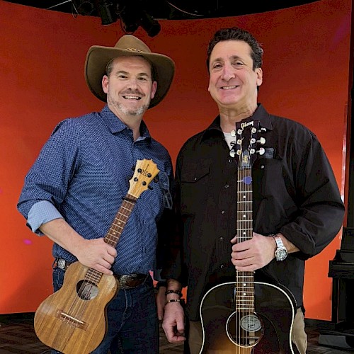 Two men with guitars, smiling on a stage with a red background, appear to be musicians ready to perform or record a show.