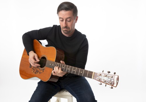 A man in a black shirt plays an acoustic guitar against a plain white background.