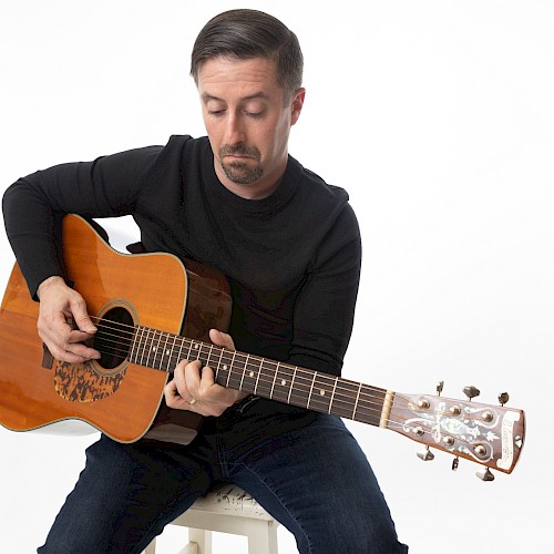 A man in a black shirt plays an acoustic guitar against a plain white background.