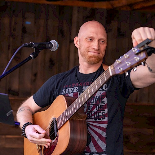 A man with a shaved head and beard is playing an acoustic guitar on stage, with a microphone nearby and a wooden background.