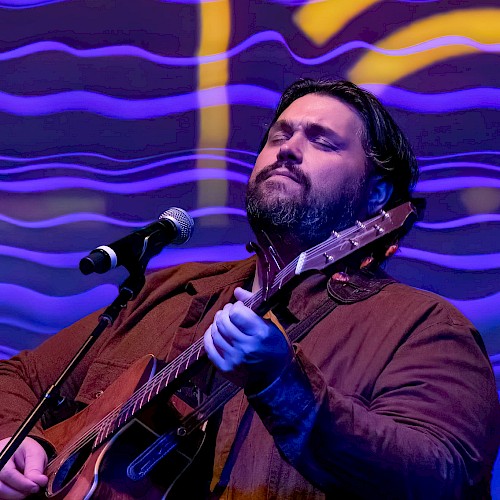 A man is serenading with a guitar and singing passionately against a vibrant, colorful, wavy background.