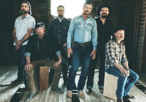 A group of six men, some sitting and some standing, pose in a rustic wooden setting, wearing casual clothes.