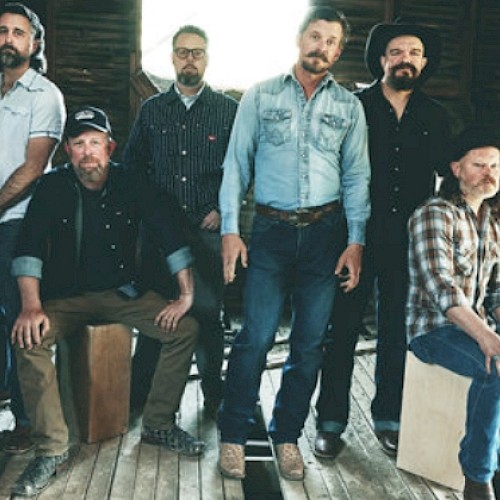 A group of six men, some sitting and some standing, pose in a rustic wooden setting, wearing casual clothes.