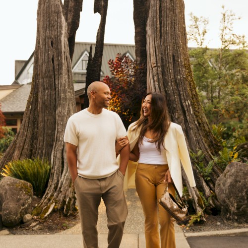 A smiling man and woman walk arm in arm on a path, with large trees and a house in the background, enjoying each other's company.