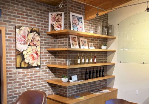 Decorative floral paintings, shelves with framed photos, bottles of wine, and a brick wall interior of a cozy cafe or wine shop.