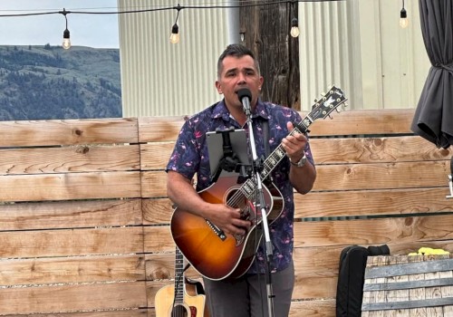 A man is performing music outdoors with a guitar and microphone, surrounded by wood fencing and mountains in the background.