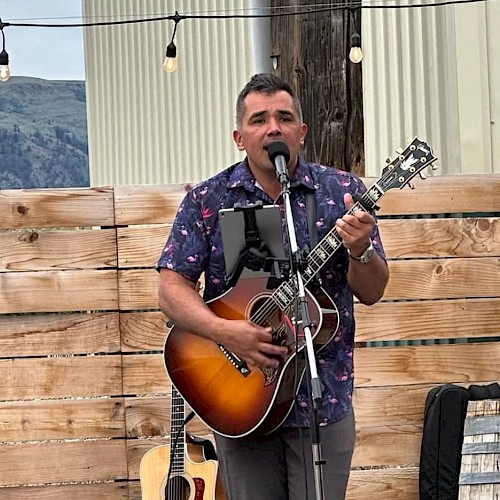 A man is performing music outdoors with a guitar and microphone, surrounded by wood fencing and mountains in the background.