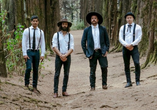 Four men dressed in vintage-style clothing stand in a forest, surrounded by tall trees and greenery.