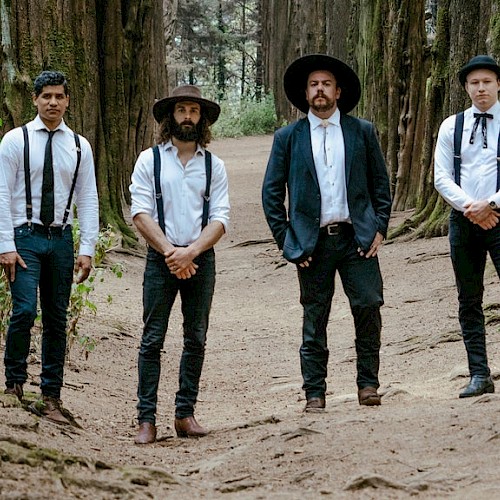 Four men dressed in vintage-style clothing stand in a forest, surrounded by tall trees and greenery.
