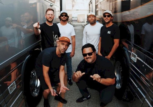 A group of six men pose between two large tour buses, some making hand gestures, in an urban alleyway, on a sunny day.