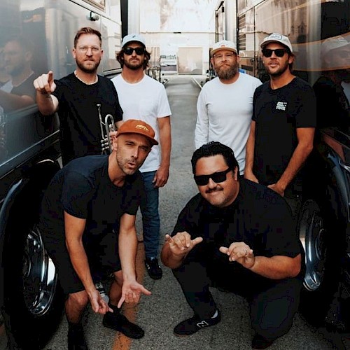 A group of six men pose between two large tour buses, some making hand gestures, in an urban alleyway, on a sunny day.