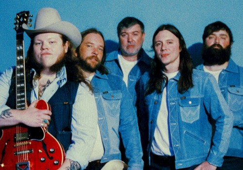 A group of five men, some with long hair and beards, pose against a blue background, with one holding a guitar and wearing a cowboy hat.
