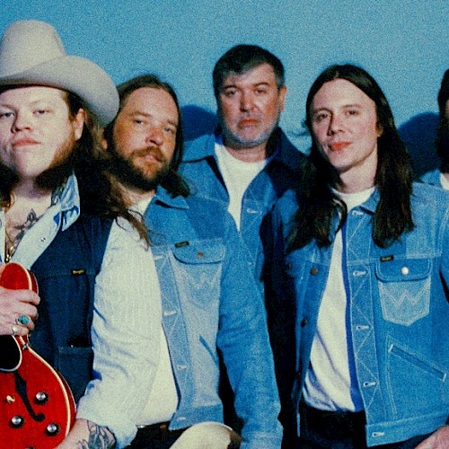 A group of five men, some with long hair and beards, pose against a blue background, with one holding a guitar and wearing a cowboy hat.