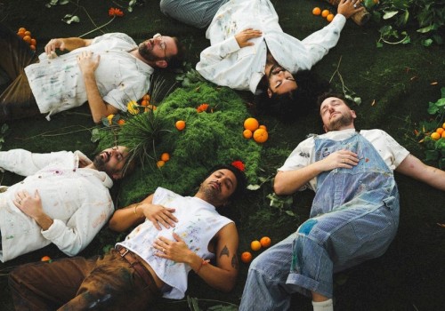 Six people lie on the ground, surrounded by greenery and oranges, on their backs with eyes closed, in a relaxed, artistic arrangement.
