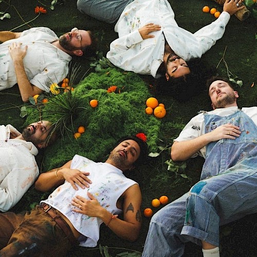 Six people lie on the ground, surrounded by greenery and oranges, on their backs with eyes closed, in a relaxed, artistic arrangement.