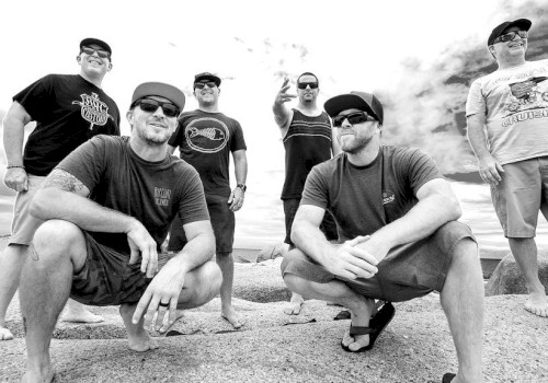 Six men are posing on a beach, some squatting and some standing, under a cloudy sky.