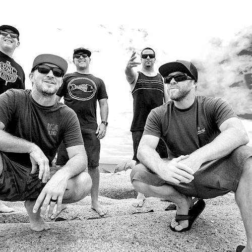 Six men are posing on a beach, some squatting and some standing, under a cloudy sky.