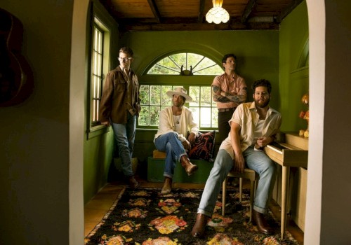 Four men in a cozy, green-walled room with a window, a guitar, and a vintage rug, creating a relaxed, artistic atmosphere.