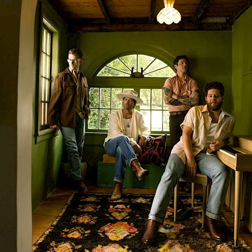 Four men in a cozy, green-walled room with a window, a guitar, and a vintage rug, creating a relaxed, artistic atmosphere.
